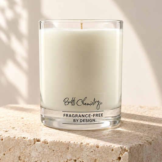 A clear glass jar candle with white wax sits on a textured stone surface against a warm beige wall, with soft shadow patterns cast across the scene. The label reads 'Buff Chemistry' and 'FRAGRANCE-FREE BY DESIGN.