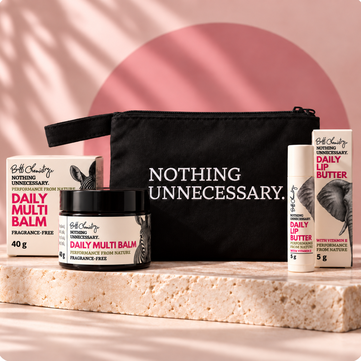 A beauty gift set featuring a black vanity zip bag, a jar of Daily Multi Balm and a tube of Daily Lip Butter arranged on a stone slab against a pink background.