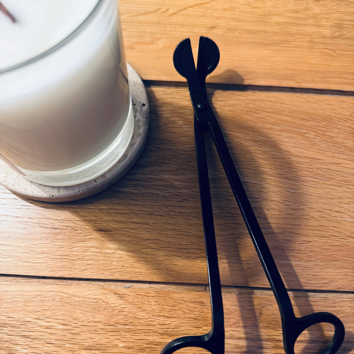 A pair of black metal wick trimmers with looped handles rests on a light brown wooden tabletop next to a white candle in a glass jar.