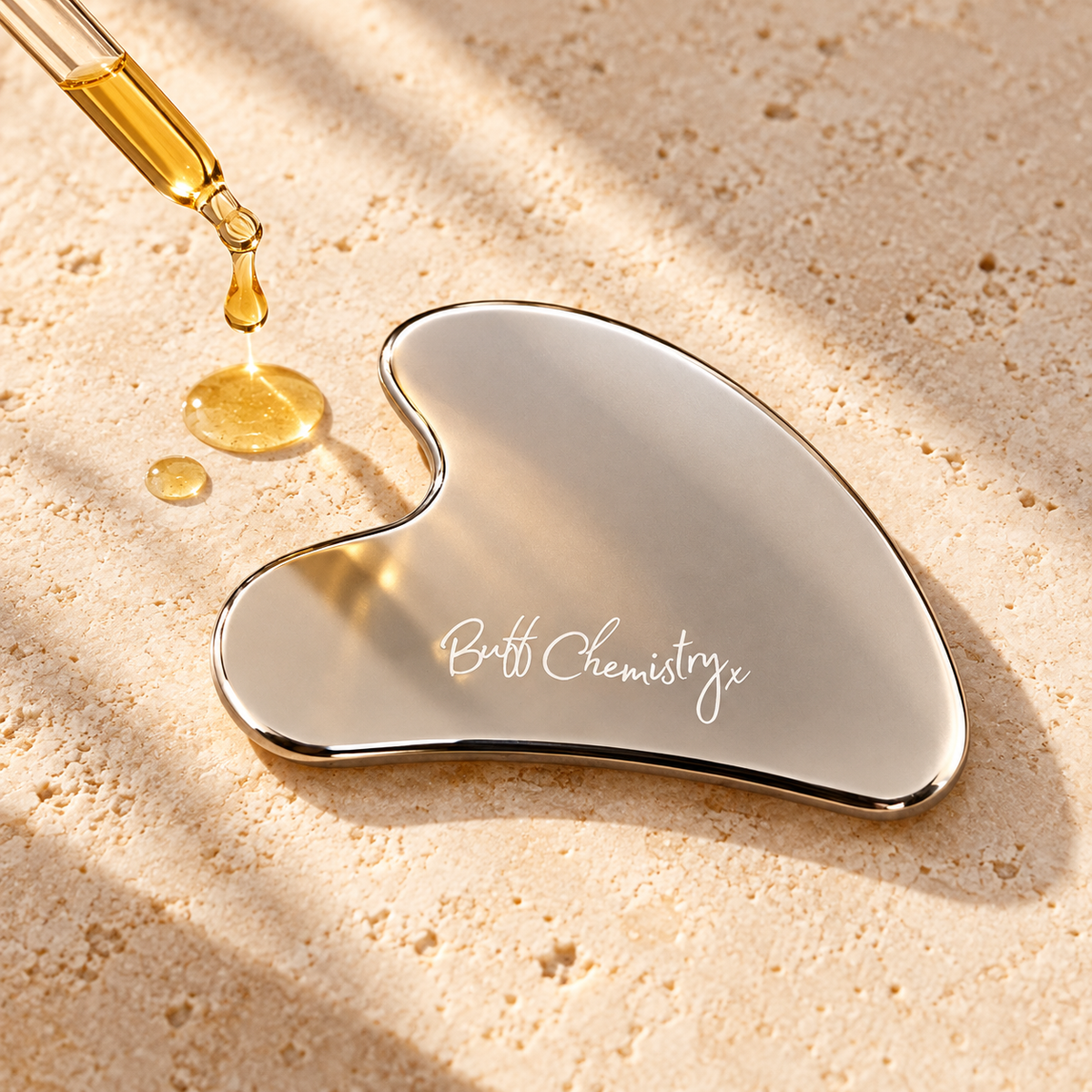 A heart-shaped stainless steel gua sha massage tool with the 'Buff Chemistry' brand name engraved on it, resting on a textured beige stone surface with a glass dropper pouring golden oil nearby.