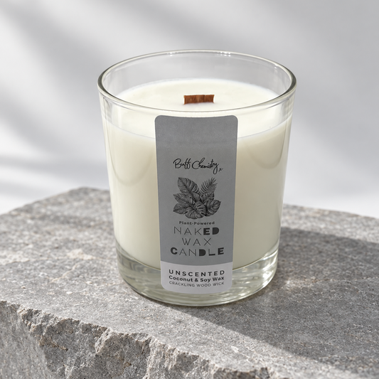 A clear glass jar candle with white wax and a small wooden wick, featuring a light blue label that reads 'Buff Chemistry NAKED WAX CANDLE' and 'UNSCENTED COCONUT & SOY WAX.' The candle sits on a gray stone slab against a white background with soft shadow overlays.