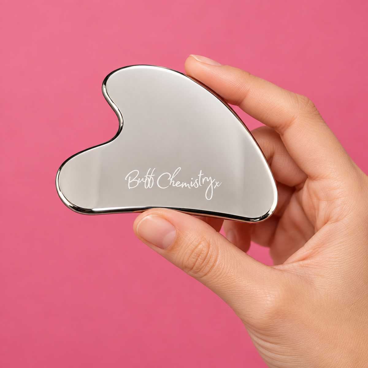 A hand holding a polished silver metal gua sha tool with the brand name 'Buff Chemistry' engraved on it, set against a solid pink background.